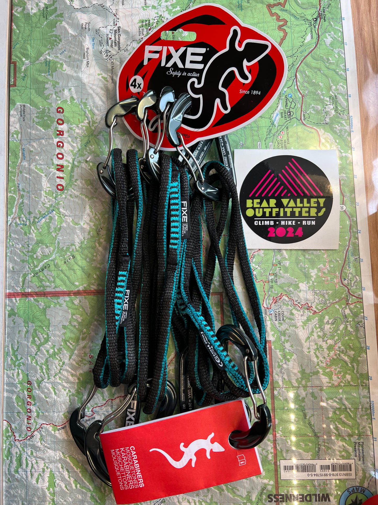 A set of four turquoise colored 60cm alpine draw carabiners with a minor bent gate design, attached to a red Fixe tag, against a background of a map or compass.